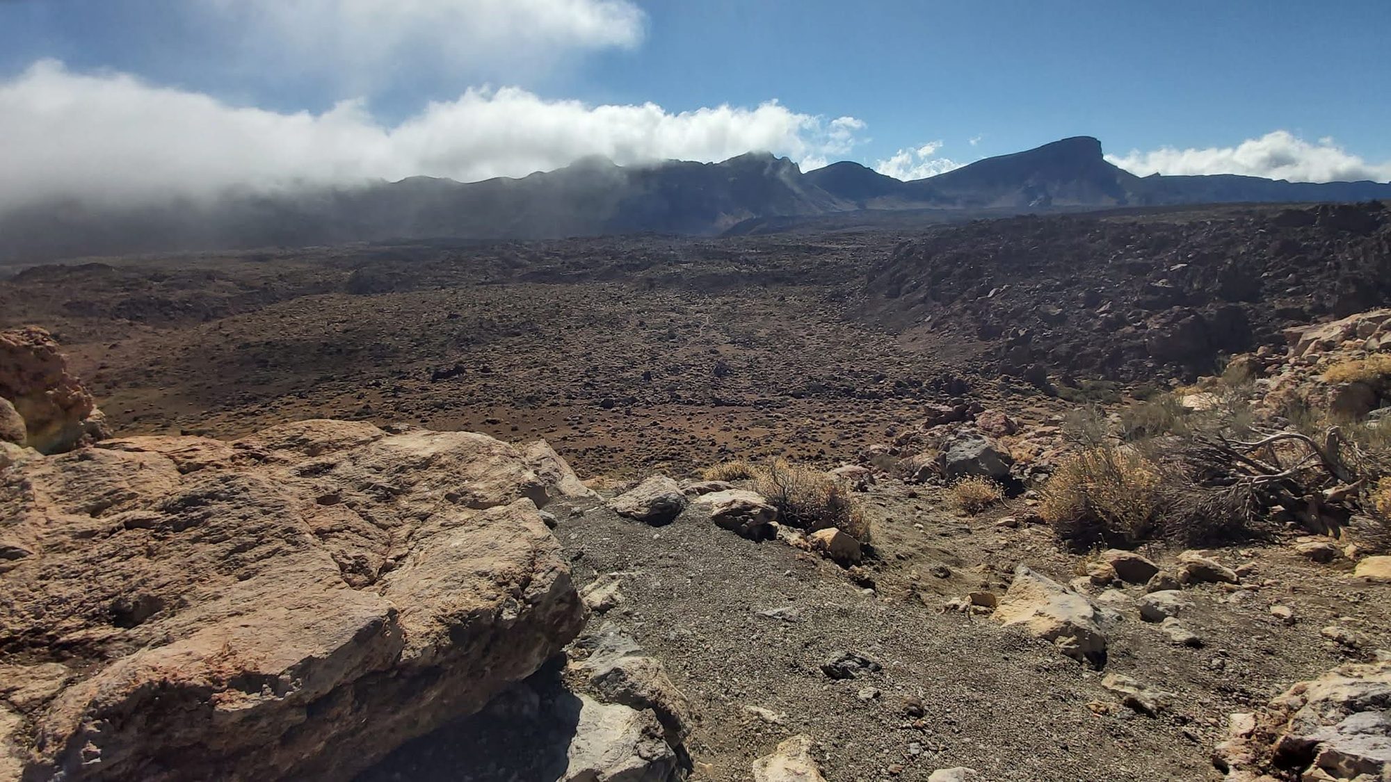 Paisaje volcánico árido y rocoso con montañas y nubes al fondo.